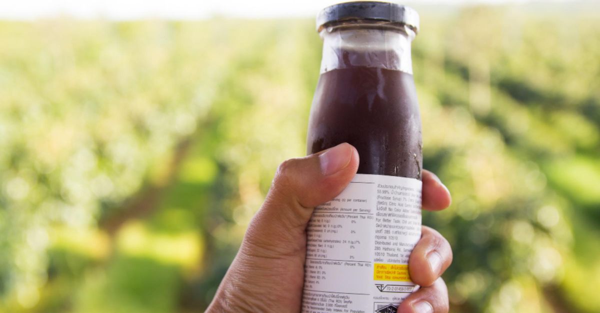 Hand holding wellness drink bottle with ingredient label in farm background.